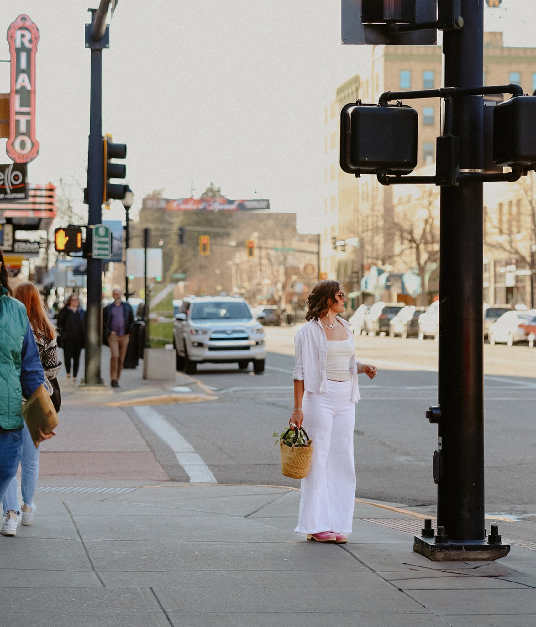 Woman in white outfit standing on a city street corner with traffic lights and pedestrians in the background.